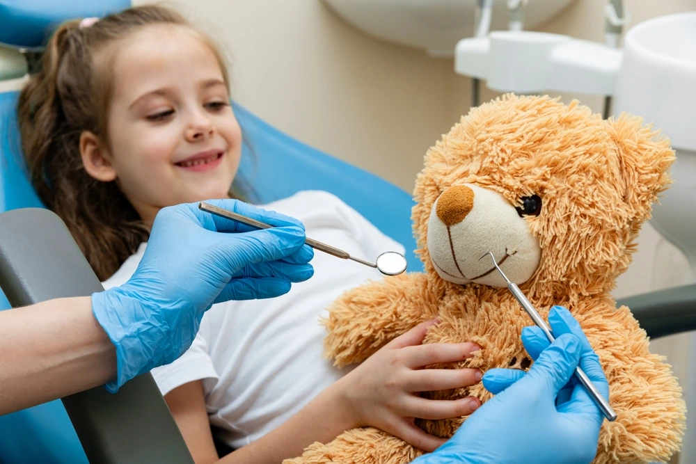A young female patient of City Family Dental Care is watching her teddy bear while her dentist is treating it